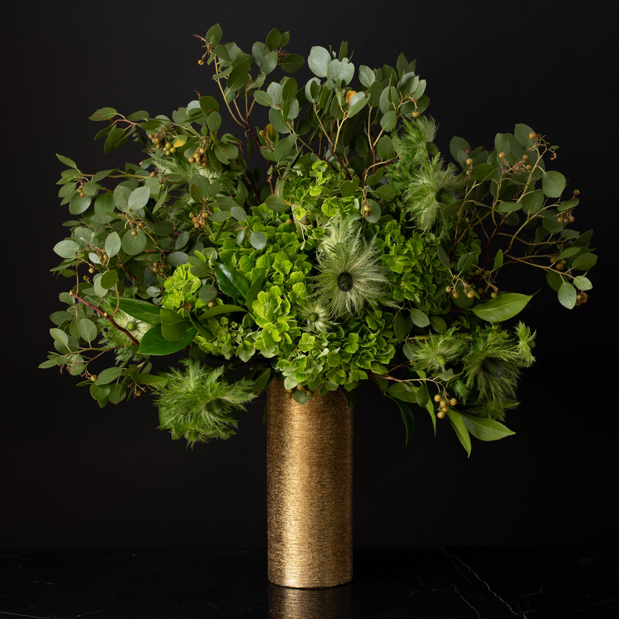 A dramatic arrangement of lush greenery, hydrangeas, and sculptural florals in a tall gold vase, designed by Bloom by Anuschka, a premier florist shop in Denver.