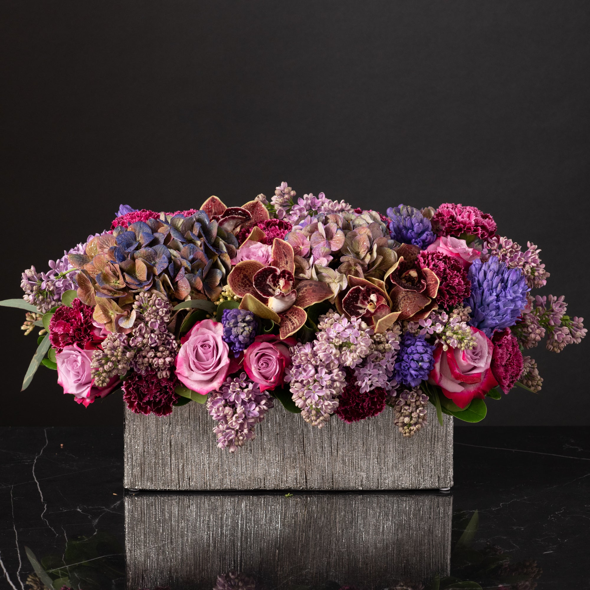 A regal long floral centerpiece featuring orchids, lilacs, roses, and hydrangeas in a textured stone vase, designed by Bloom by Anuschka, a premier florist shop in Denver.